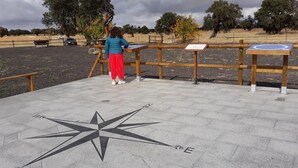 A person standing at a wooden observation post with a compass on the ground, surrounded by a park with trees and a car in the background. - Pension Ruda (Hinojosa del Duque)