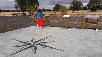 Una persona de pie en un puesto de observación de madera con una brújula en el suelo, rodeada por un parque con árboles y un coche al fondo.
