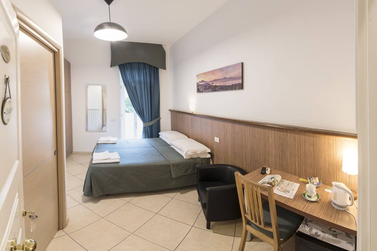 A hotel room with a bed, a desk, a chair, a small table, a wall-mounted painting, and a door.