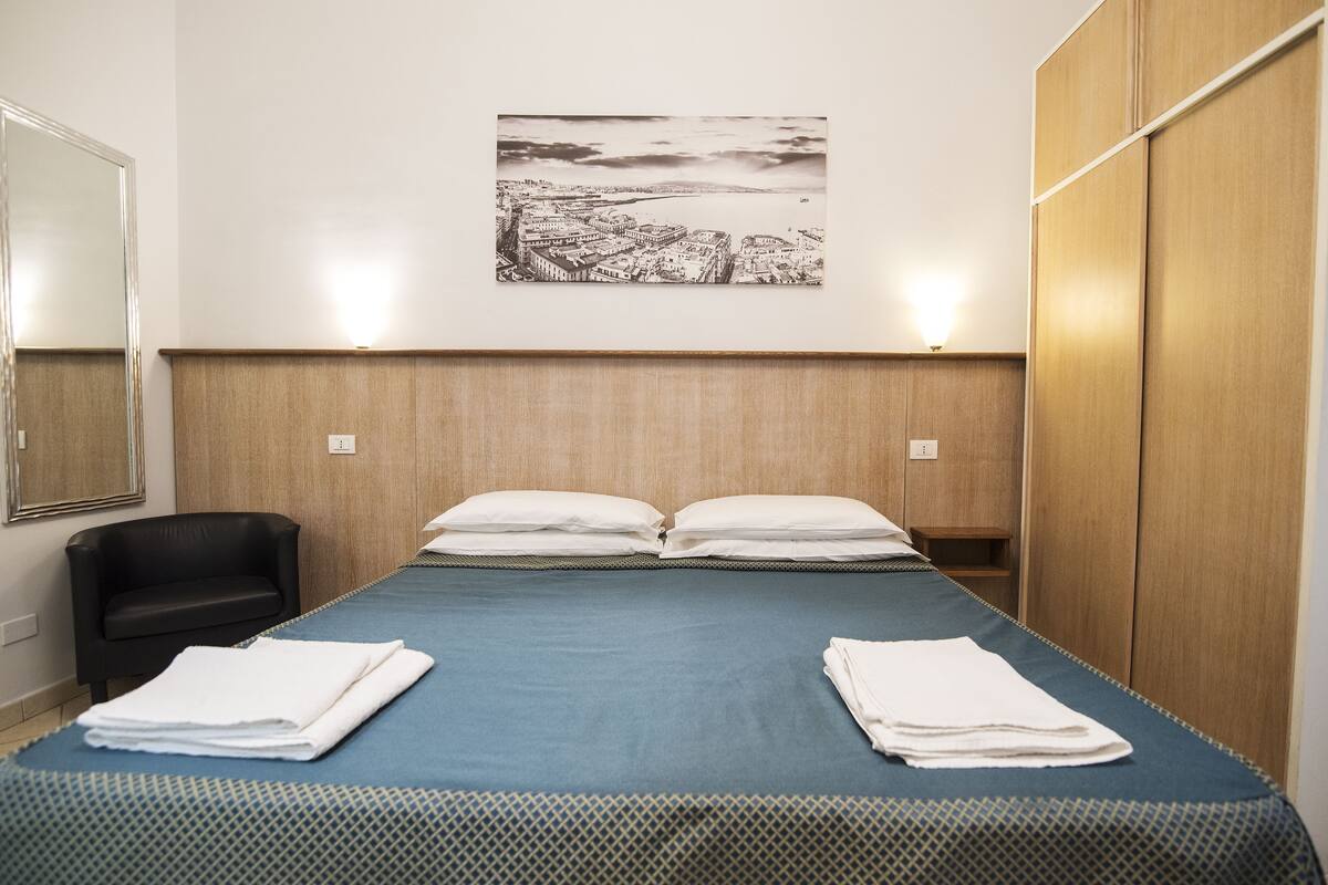 A hotel room with a bed, two pillows, a chair, a wardrobe, and a wall-mounted picture.