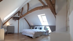 Een slaapkamer met een schuine plafond, houten balken, een bed met witte beddengoed, een dressoir, een spiegel en een raam.