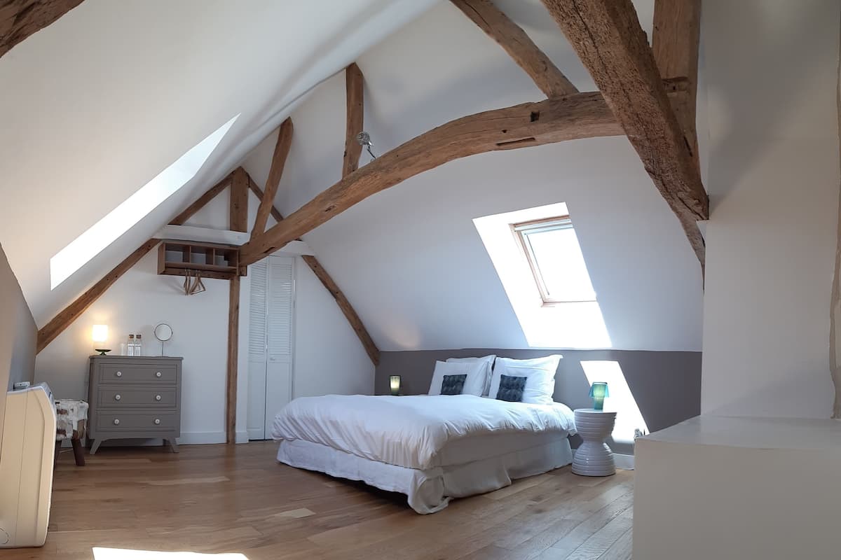 Une chambre avec un plafond en pente, des poutres en bois, un lit avec une literie blanche, une commode, un miroir et une fenêtre.