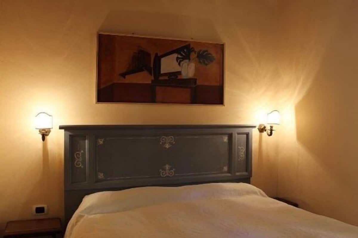 Un dormitorio con cabecera oscura, un cuadro sobre la cama y dos lámparas fijadas a la pared.