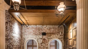 Una habitación acogedora con paredes de ladrillo visto, vigas de madera y un gran ventanal arqueado. Se observan dos bancos de madera y una mesa con sillas.