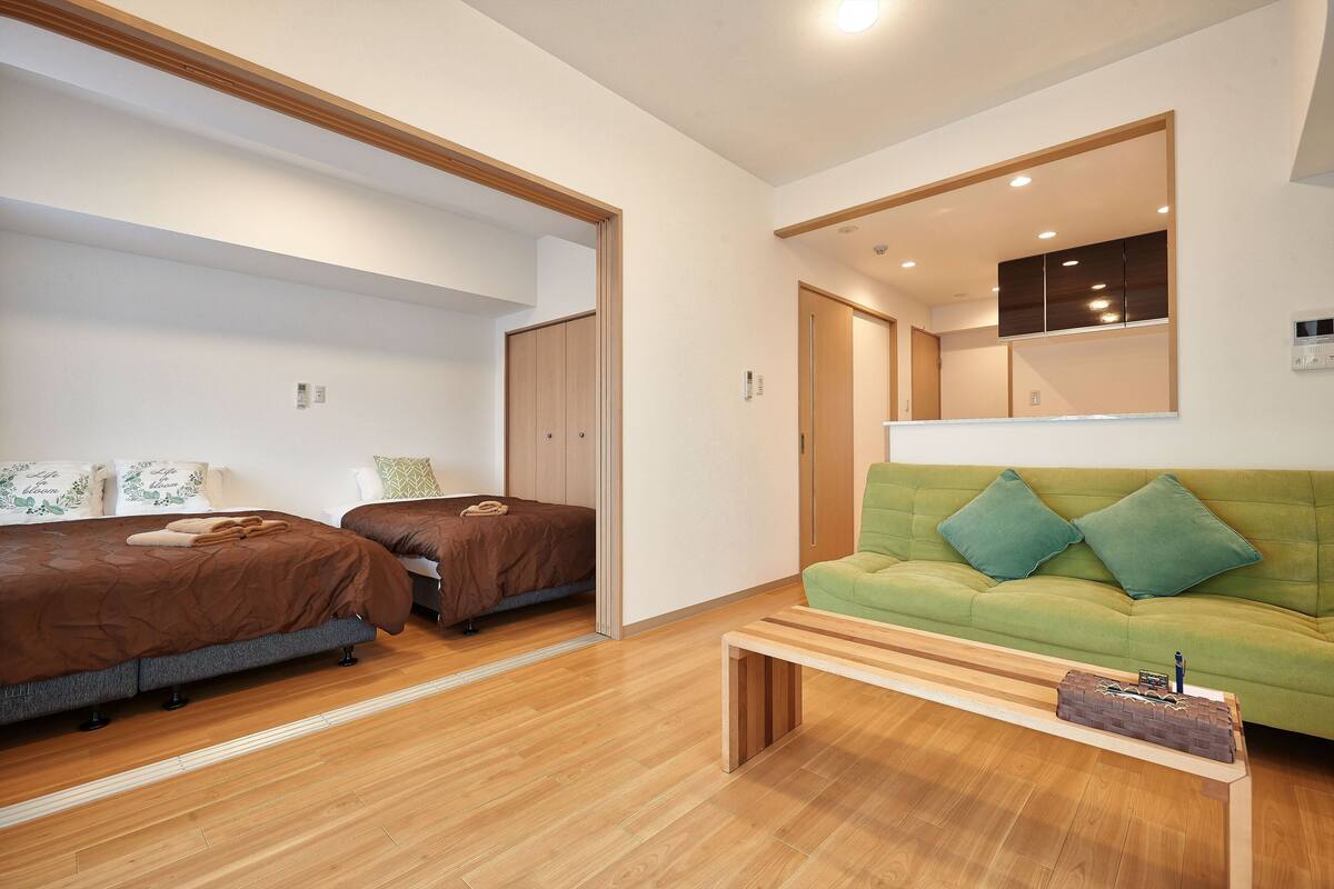 Quarto de hotel moderno com piso de madeira, um sofá verde com duas almofadas, uma mesa de centro de madeira e duas camas com roupa de cama marrom.