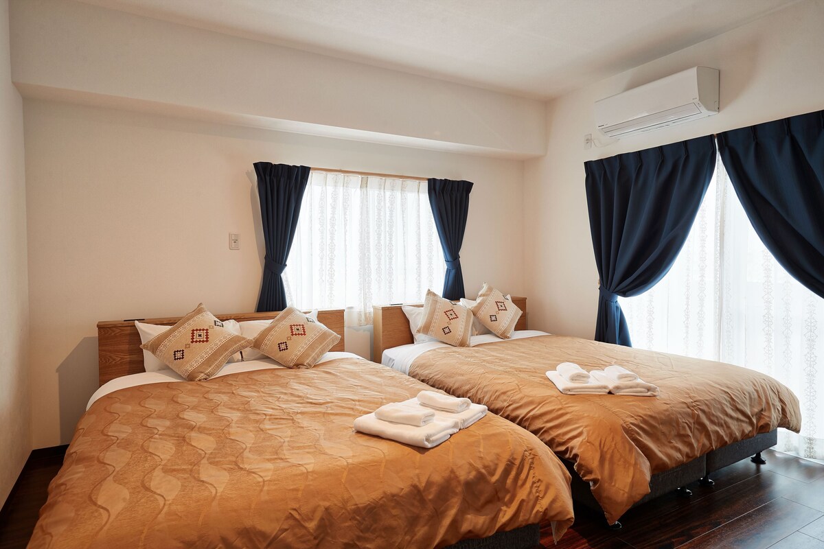 A hotel room with two beds, each with a brown bedspread and matching pillows. There is an air conditioning unit on the wall and a window with dark curtains.
