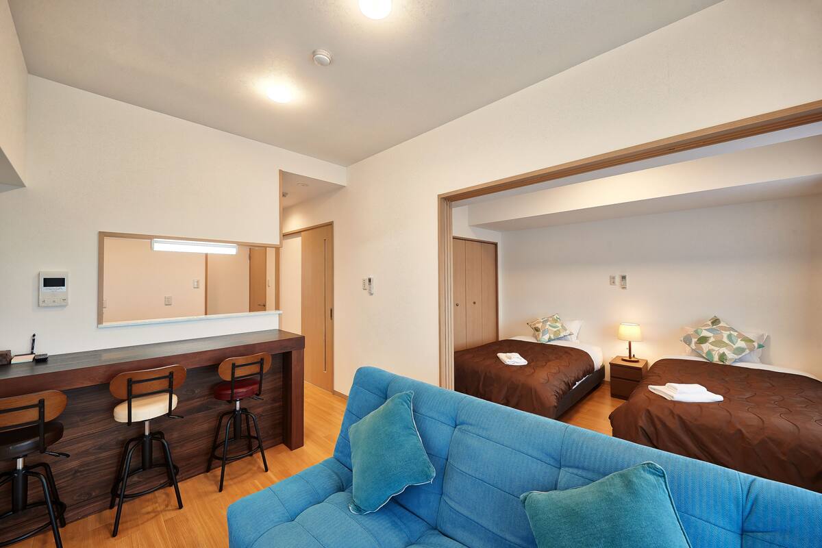 Quarto de hotel com sofá azul, duas camas de solteiro, uma escrivaninha com cadeiras e um espelho sobre a escrivaninha.