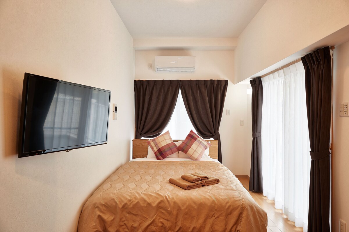 A hotel room with a bed, a flat-screen TV, a window with curtains, and an air conditioning unit.