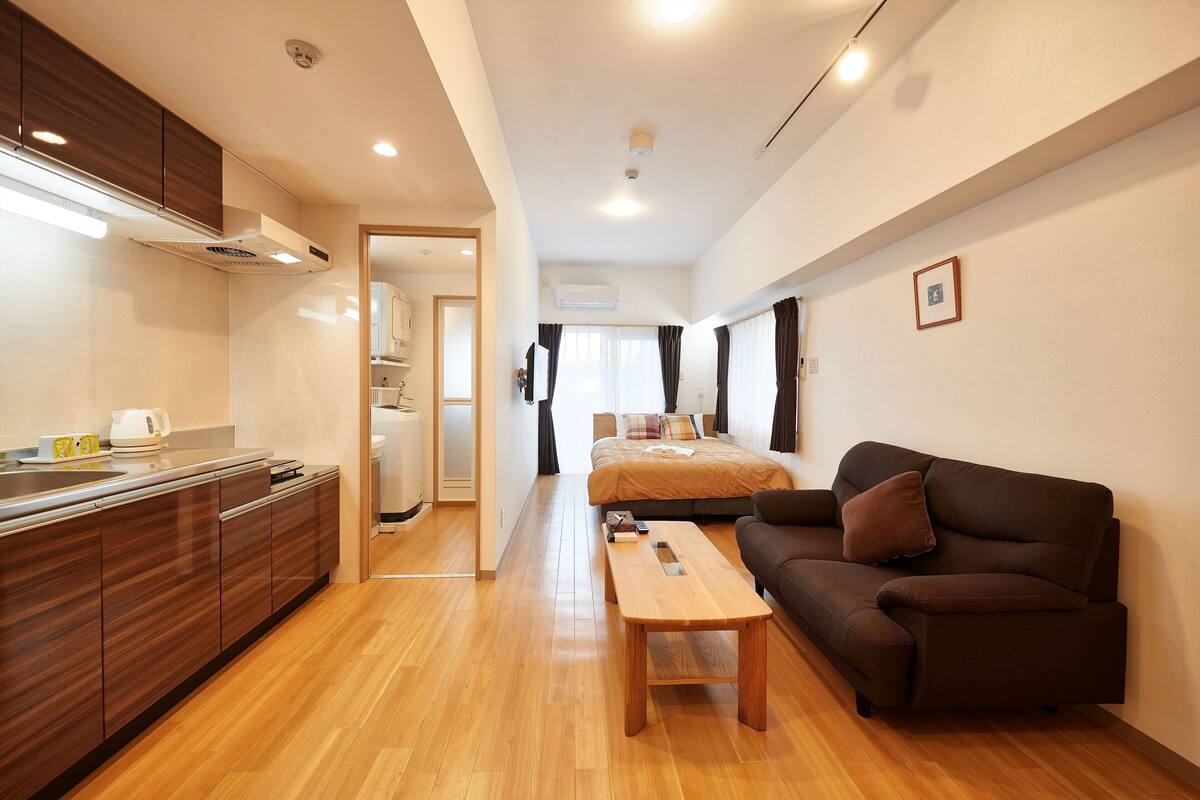 Quarto de hotel moderno com piso de madeira, sofá, mesa de madeira, área de cozinha com armários e pia, e uma cama grande com vista para o exterior.