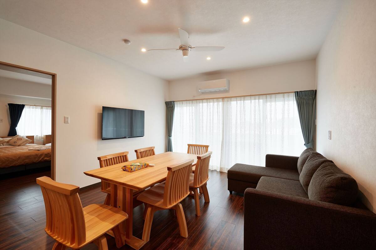 Quarto moderno com mesa de jantar de madeira, cadeiras, sofá, ventoinha no teto, televisão e uma janela grande com cortinas.