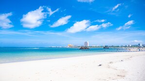 Una playa con agua azul cristalina, arena blanca y un horizonte distante con edificios y una noria.