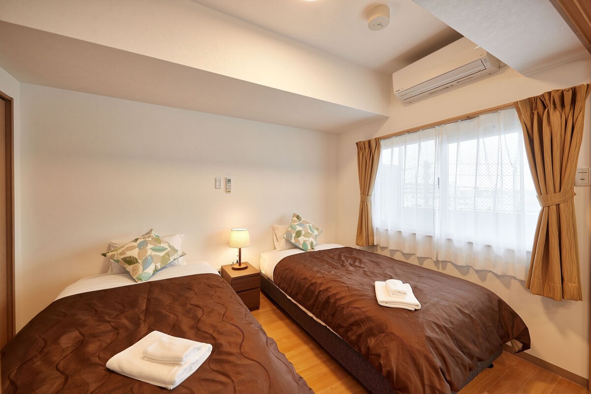 A hotel room with two beds, a nightstand with a lamp, an air conditioning unit, and a window with curtains.