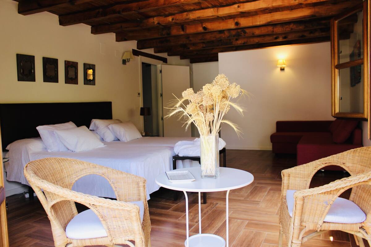 A hotel room with a bed, two wicker chairs, a small white table, and a vase with dried flowers.
