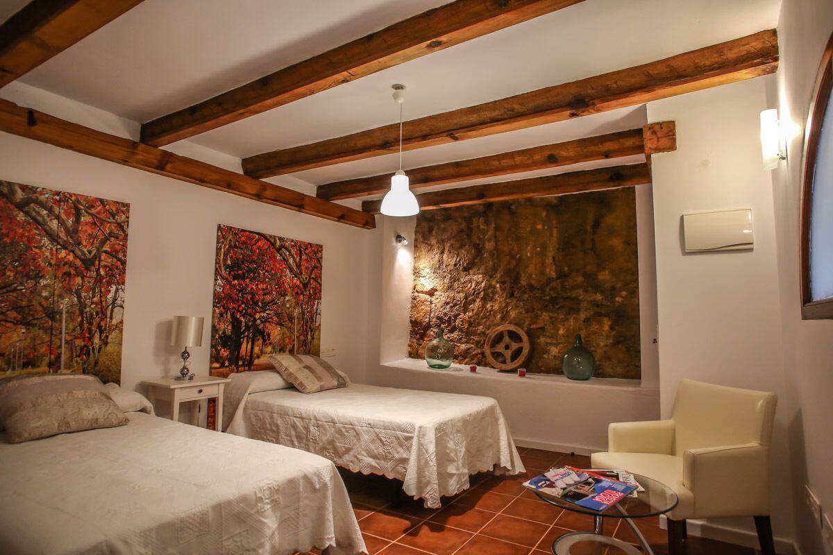 A room with two beds, a chair, a small table, and a stone wall with a window.
