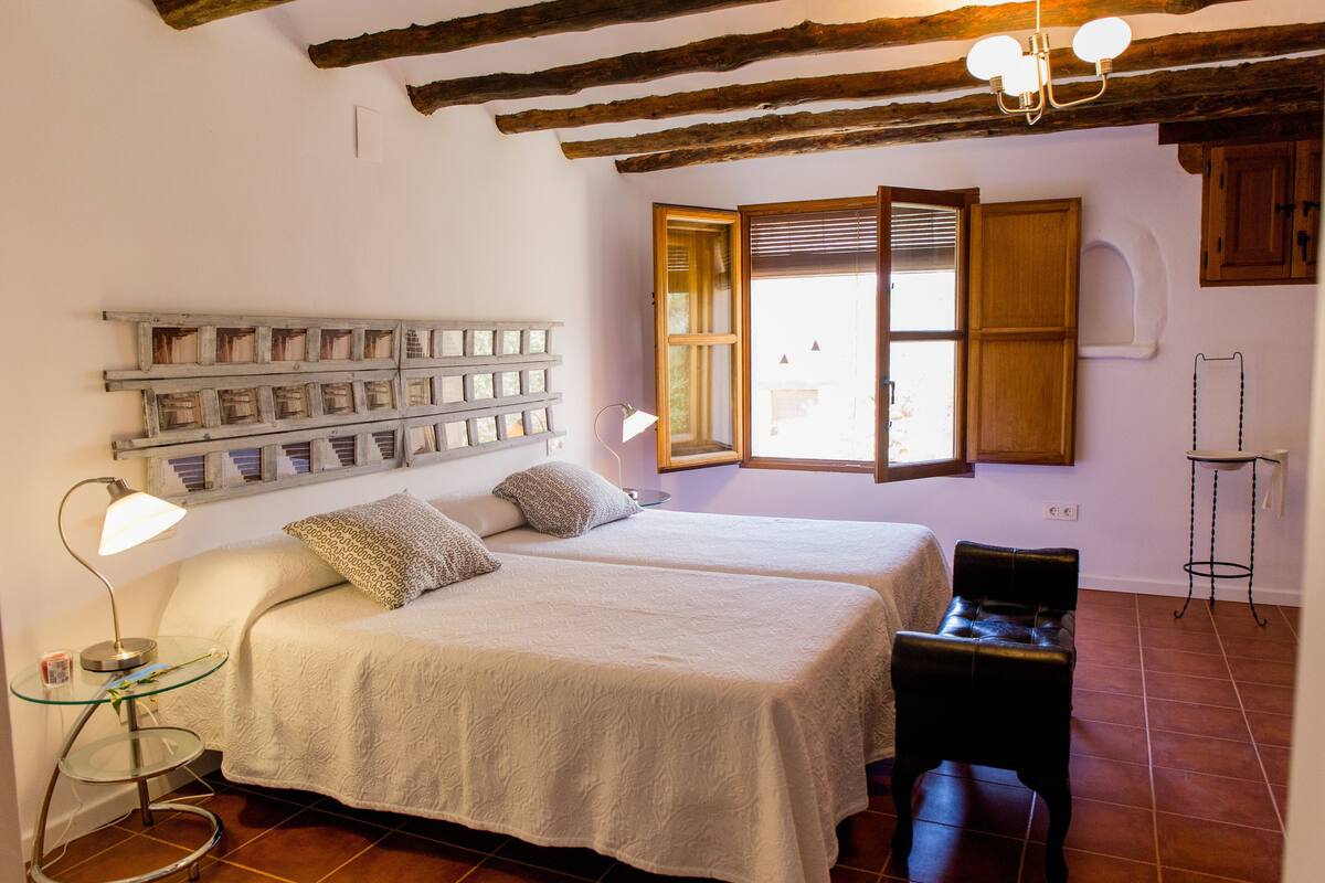 A bedroom with two beds, a wooden ceiling, a window with blinds, and a small table with a lamp.
