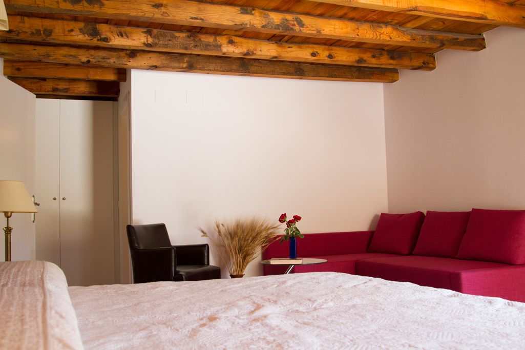 Ein Hotelzimmer mit Holzboden, einem Bett mit weißer Bettdecke, einem roten Sofa, einem schwarzen Sessel und einem kleinen Tisch mit Blumen.