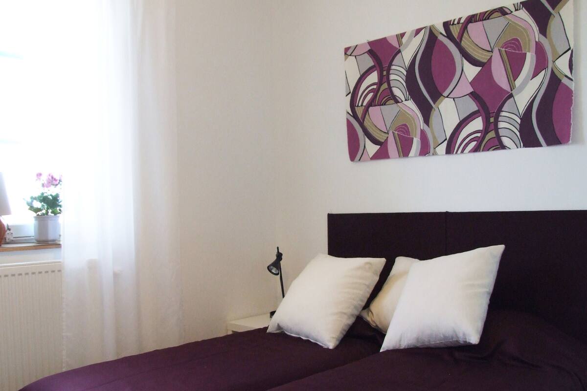 A room with a bed, two pillows, a wall-mounted artwork, and a window with curtains.
