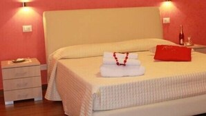 Una cama bien hecha con cubrecama blanco y almohada roja, una mesita de noche con una botella, y un objeto rojo sobre la cama.