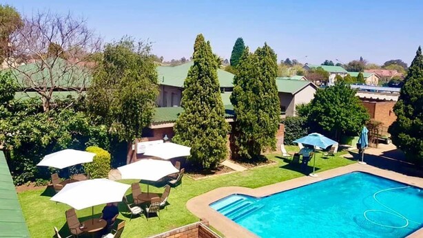 A backyard with a swimming pool, surrounded by trees and a patio area with umbrellas. - Elgro Hotel (Potchefstroom)
