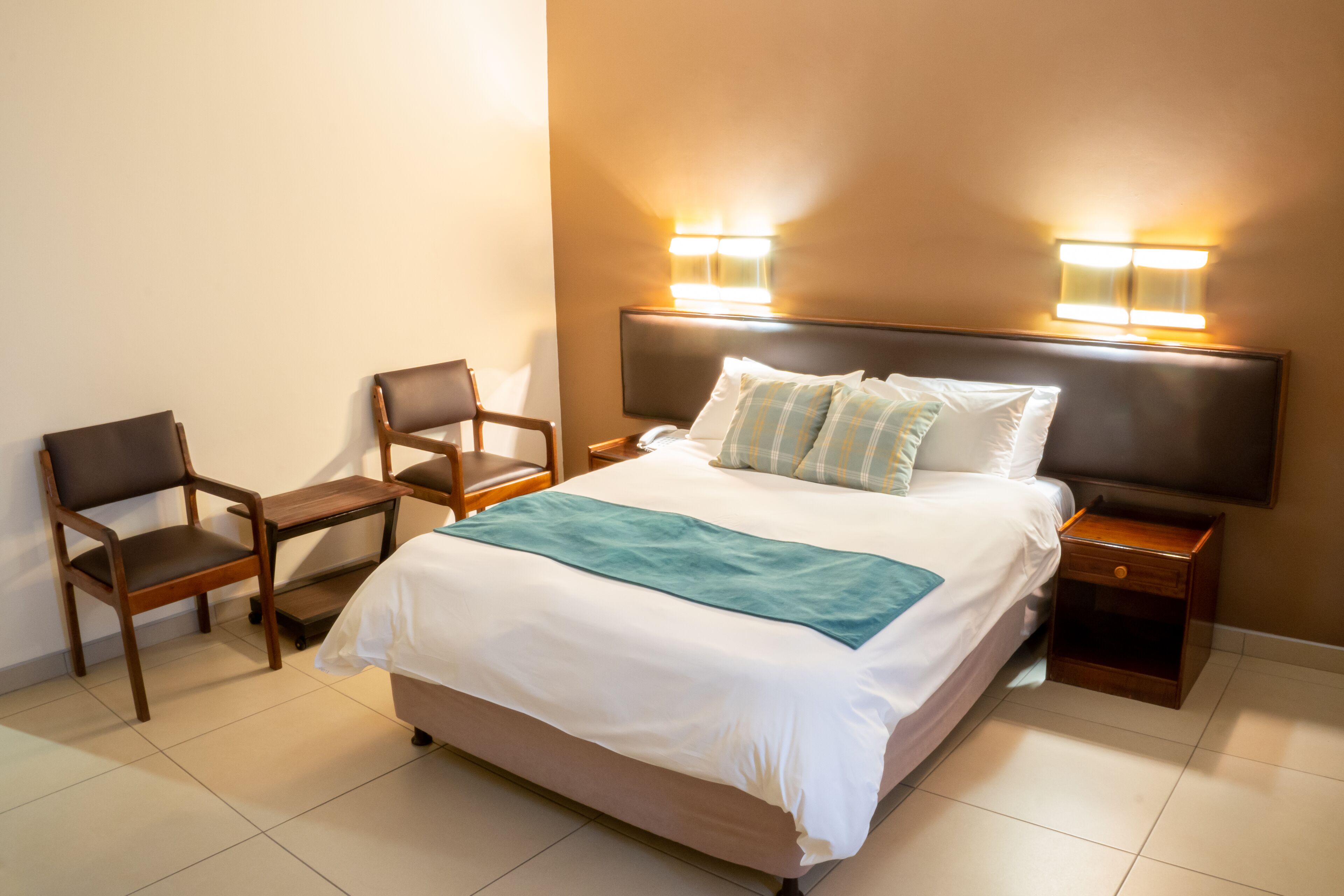 Una habitación de hotel ordenada con cama, dos sillas, una mesita de noche e iluminación en la pared.