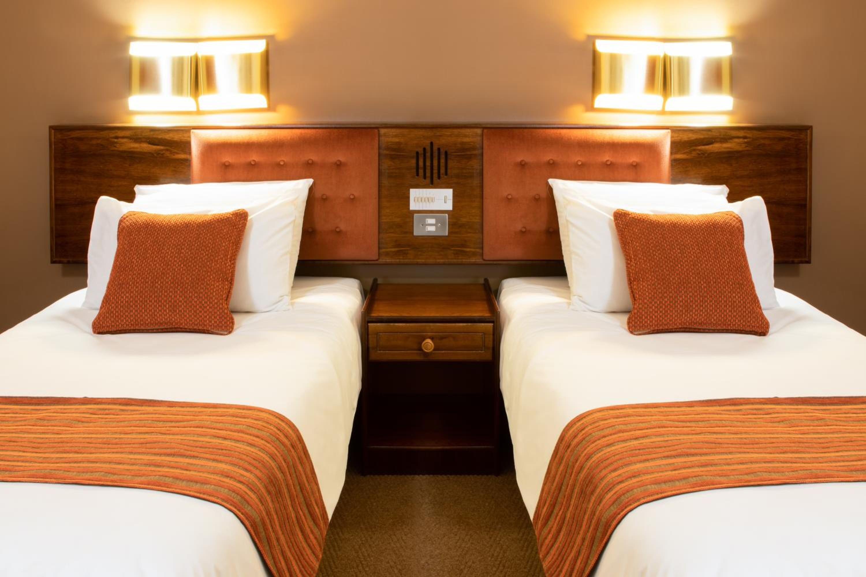 A hotel room with two beds, each with a wooden headboard, white bedding, and an orange throw pillow. There are two wall-mounted lights above each bed, and a small wooden nightstand with a drawer between the beds.