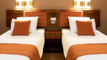 A hotel room with two beds, each with a wooden headboard, white bedding, and an orange throw pillow. There are two wall-mounted lights above each bed, and a small wooden nightstand with a drawer between the beds.