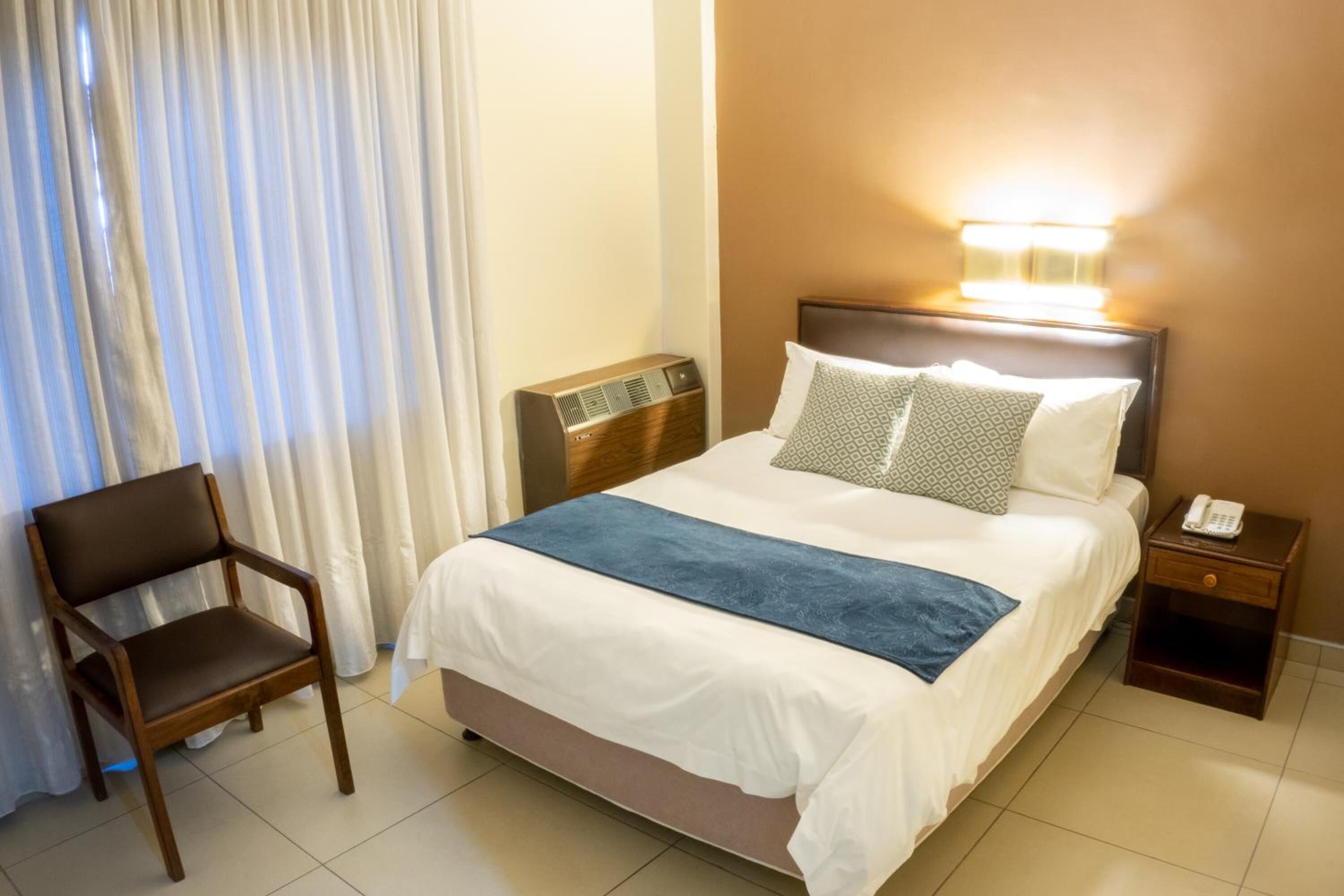 A hotel room with a bed, a chair, a bedside table with a phone, and a wall-mounted light.