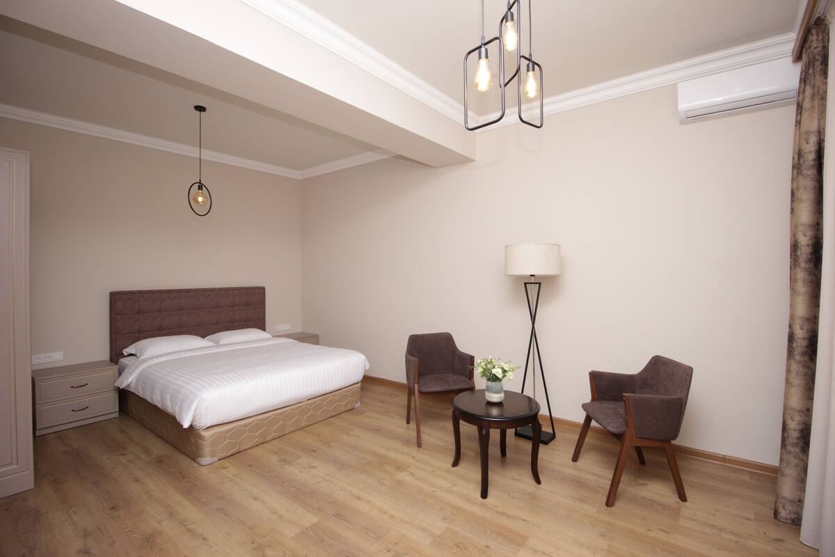 A hotel room with a bed, two armchairs, a small table with a vase, a floor lamp, and a ceiling light fixture.