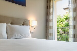 Camera d'albergo con un letto grande, una testiera, una lampada a parete e vista sul verde attraverso tende leggere.