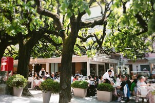 Zona pranzo all'aperto con tavoli e sedie, circondata da alberi e un edificio con un cartello.