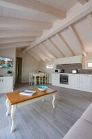 Amplia sala de estar con una mesa de comedor y sillas, una cocina con gabinetes y electrodomésticos blancos, y un techo alto con vigas a la vista.