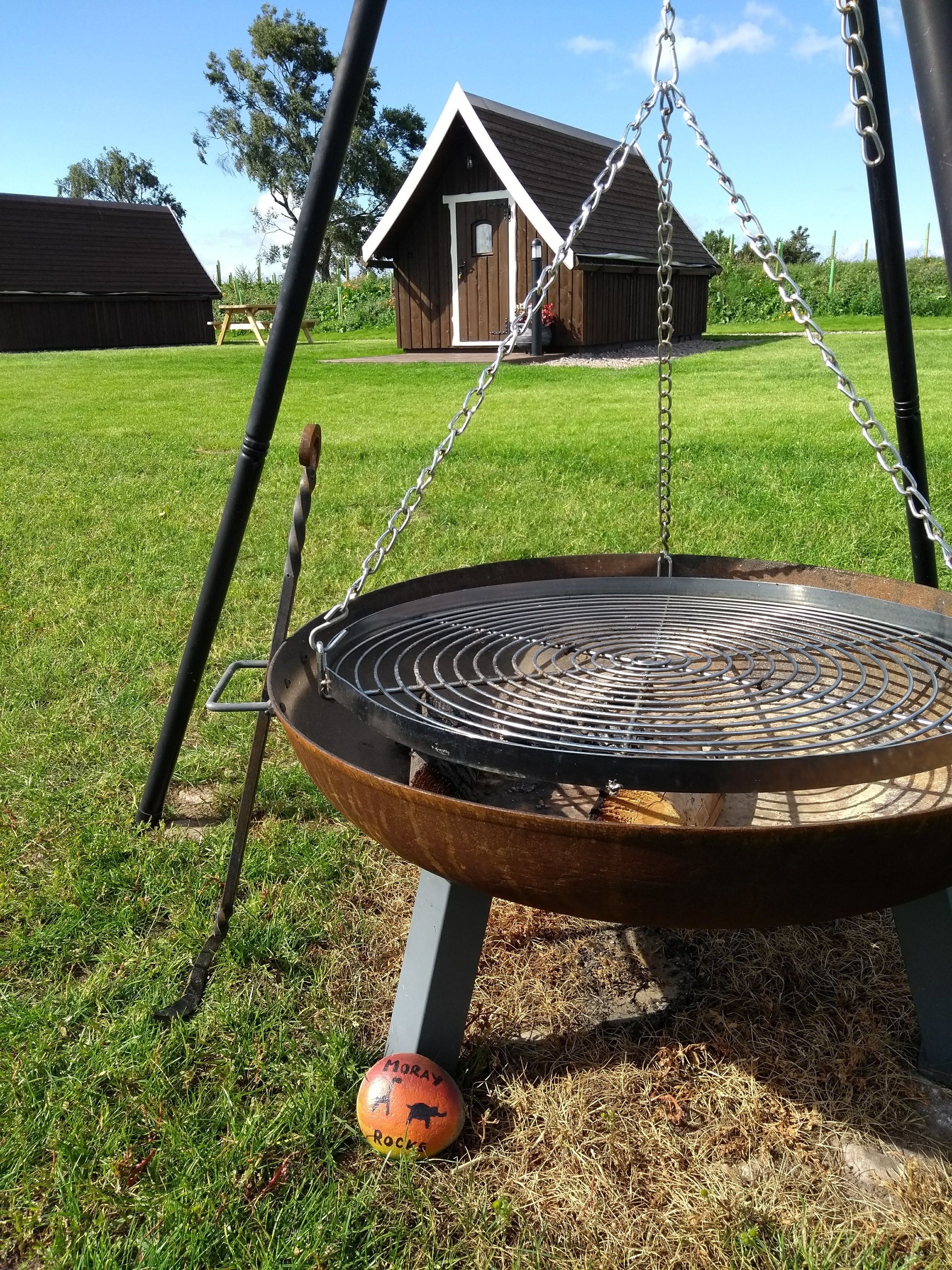 Un gril en métal avec une chaîne suspendue, placé sur une pelouse verdoyante, avec un bâtiment en bois en arrière-plan.
