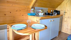 A cozy kitchen with wooden walls and a sloped ceiling. A wooden table with plates and a wine glass is set up. A sink and stove are visible in the background. - Macbeth's Hillock (Nairn)