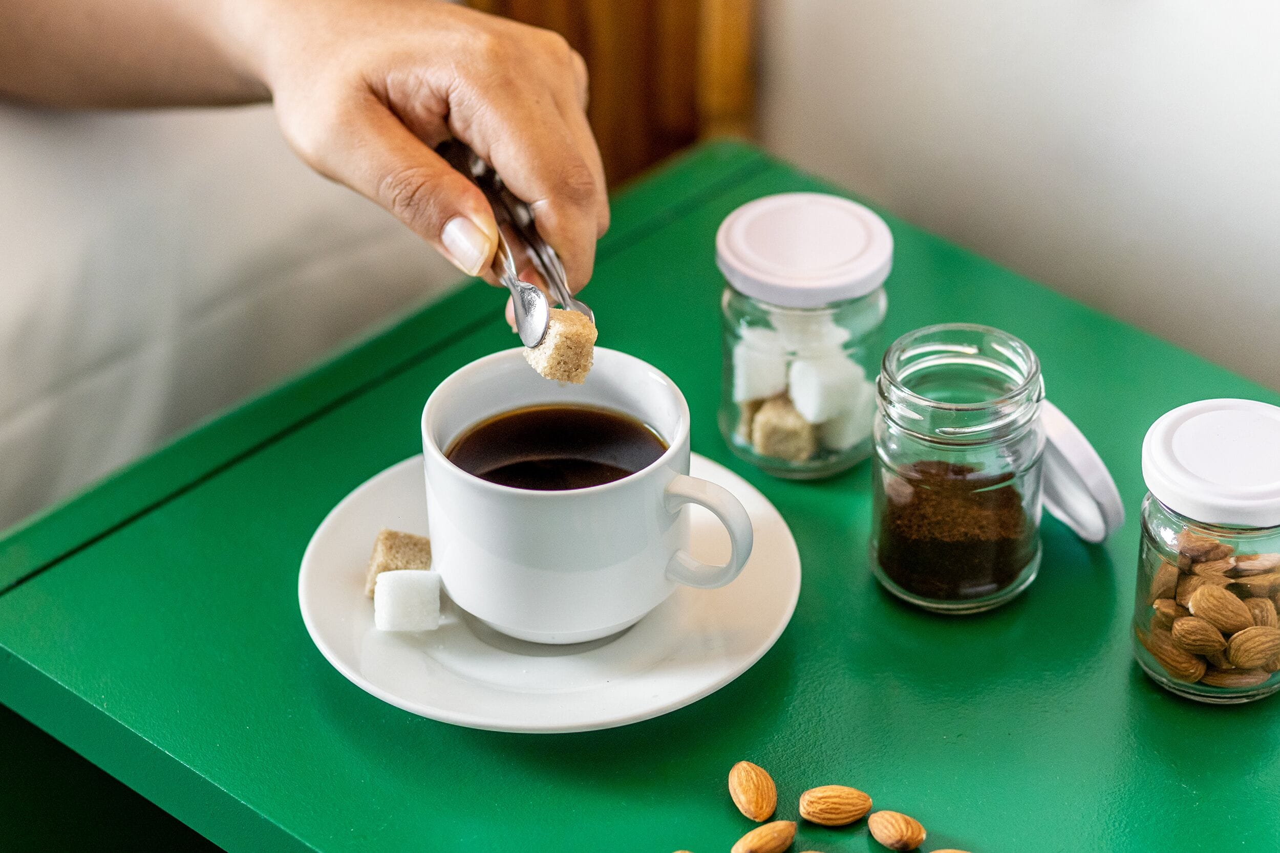 Eine weiße Kaffeetasse mit einem Löffel Zucker auf einem grünen Tablett. Zwei Vorratsgläser mit Deckeln stehen daneben, eines gefüllt mit braunem Pulver, das andere mit Mandeln.
