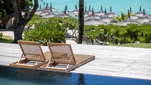 Un espace aménagé au bord de la piscine, avec deux chaises longues, une terrasse en bois et une vue sur l’océan, agrémenté de parasols en feuilles de palmier.