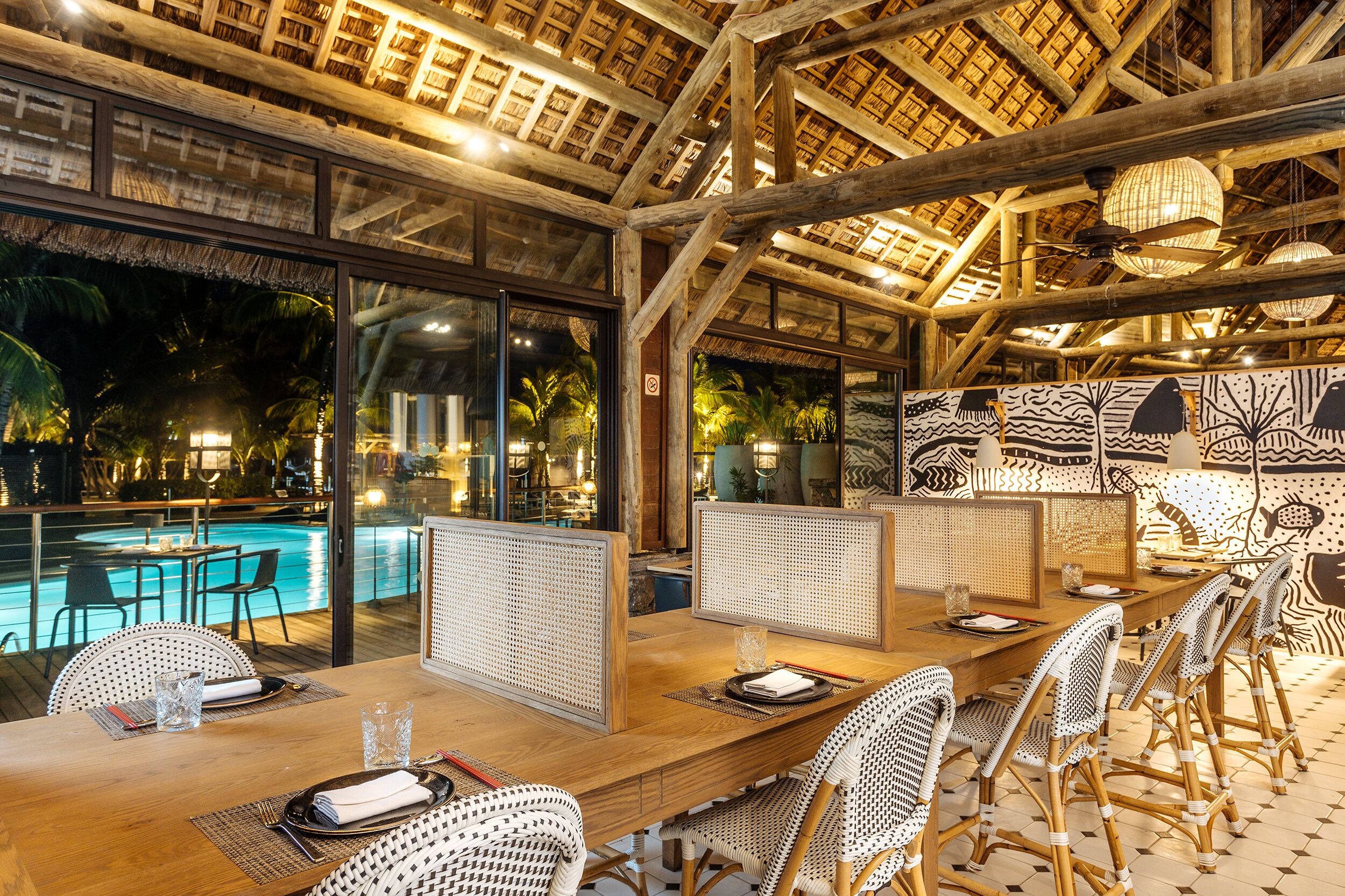Un comedor interior con vista a la piscina, vigas de madera y sillas tejidas.