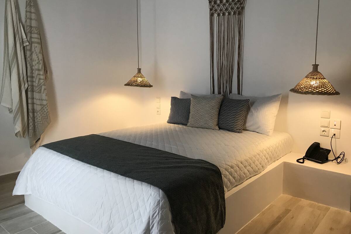 Una cama bien tendida con un colcha blanca y gris oscuro, dos tapices de macramé colgando en la pared y dos lámparas colgantes.