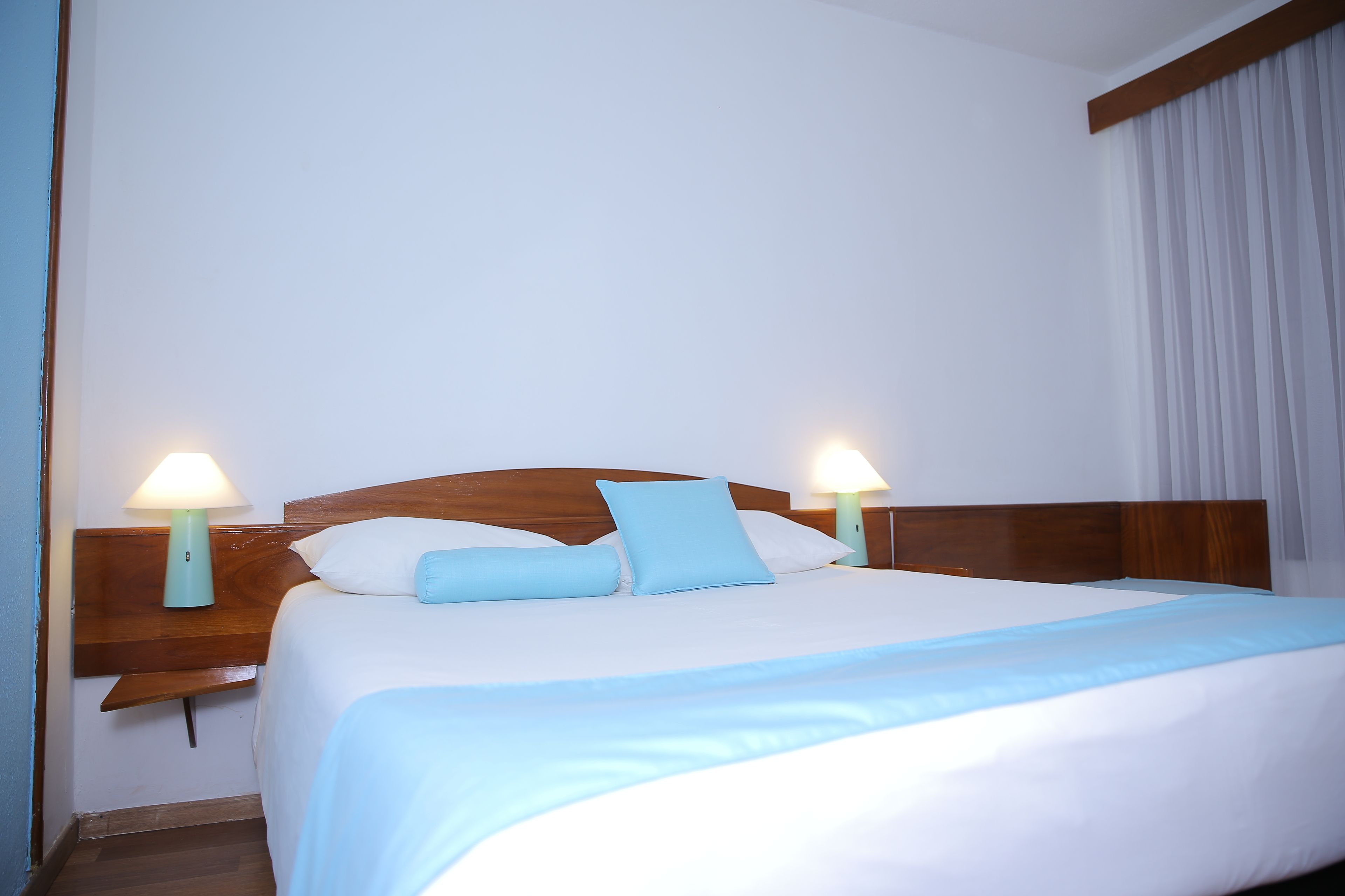 A neatly made bed with a wooden headboard, two bedside lamps, and a white curtain.