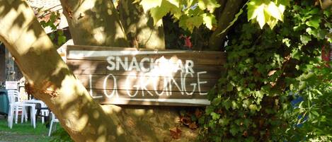A wooden sign with graffiti that reads "SNACK BAR LO CRONGE" attached to a tree.