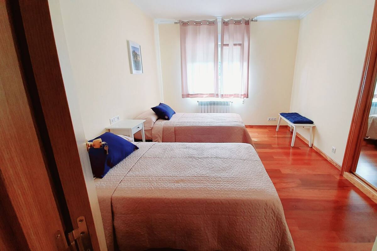 Una habitación con dos camas, una mesita pequeña, un banco, un radiador y una ventana con cortinas.