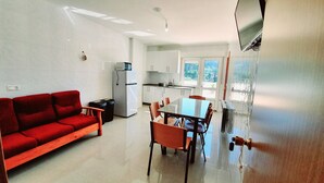 Ein kompakter Wohnbereich mit einem roten Sofa, einem Esstisch mit Stühlen, einer Küchenzeile inklusive Kühlschrank und einem an der Wand montierten Fernseher.