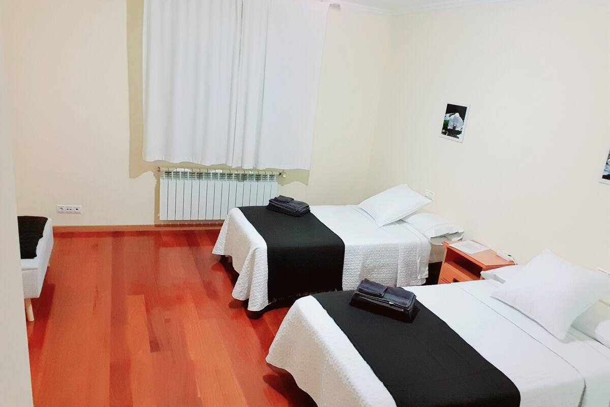 Habitación de hotel con dos camas, un radiador, una ventana con cortinas y una mesita con una silla.