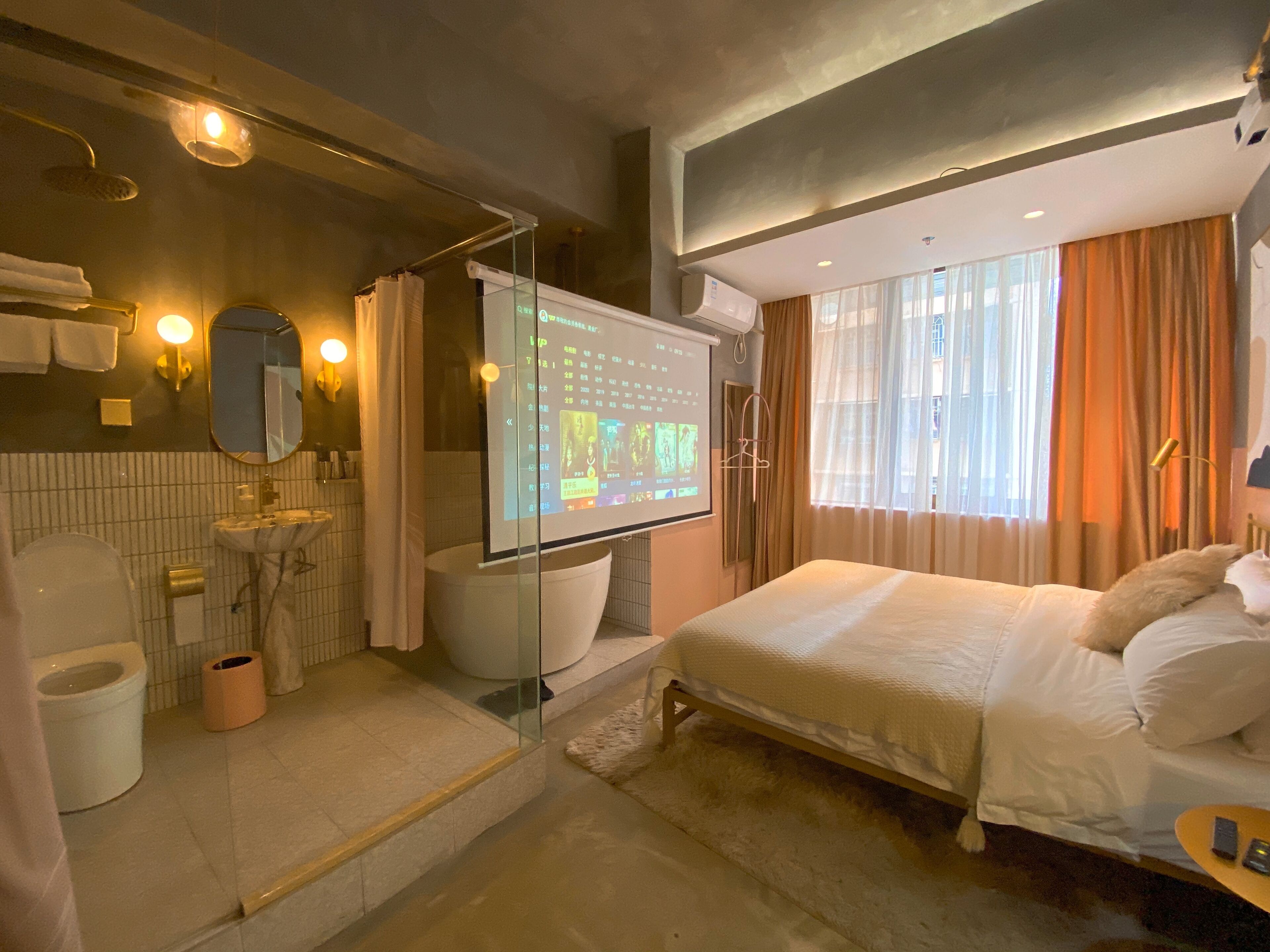 Una habitación de hotel moderna con una cama grande, baño separado con ducha, mampara de vidrio y una pantalla de proyección que muestra información.