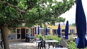 An outdoor seating area with tables and chairs, covered by blue umbrellas, and surrounded by trees. - Camping Les Sources (Courthezon)
