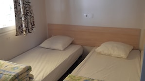 A room with two single beds, each with a pillow and a blanket. There is a window with a curtain, and a shelf above the beds. - Camping Les Sources (Courthezon)