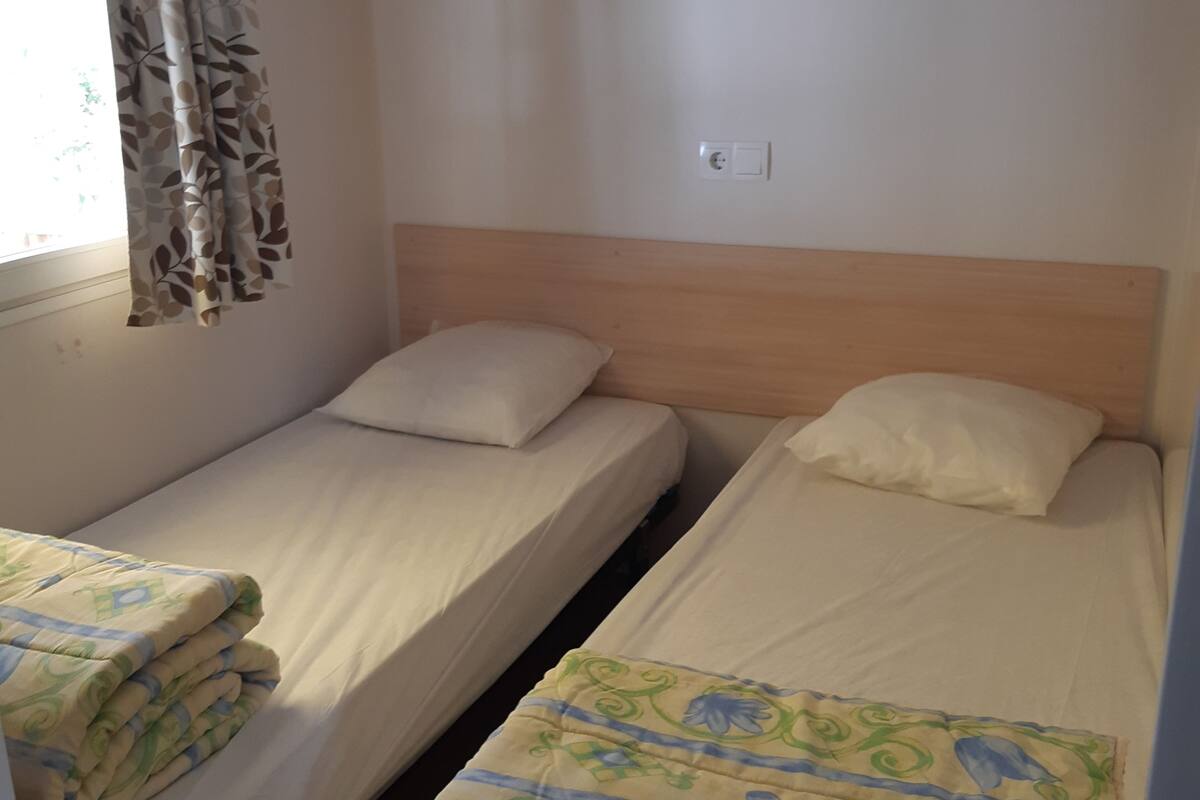 A room with two single beds, each with a pillow and a blanket. There is a window with a curtain, and a shelf above the beds.