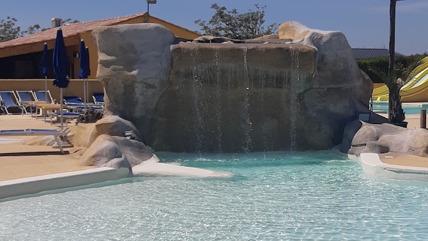 A pool with clear water, a rock formation, and lounge chairs. - Camping Les Sources (Courthezon)
