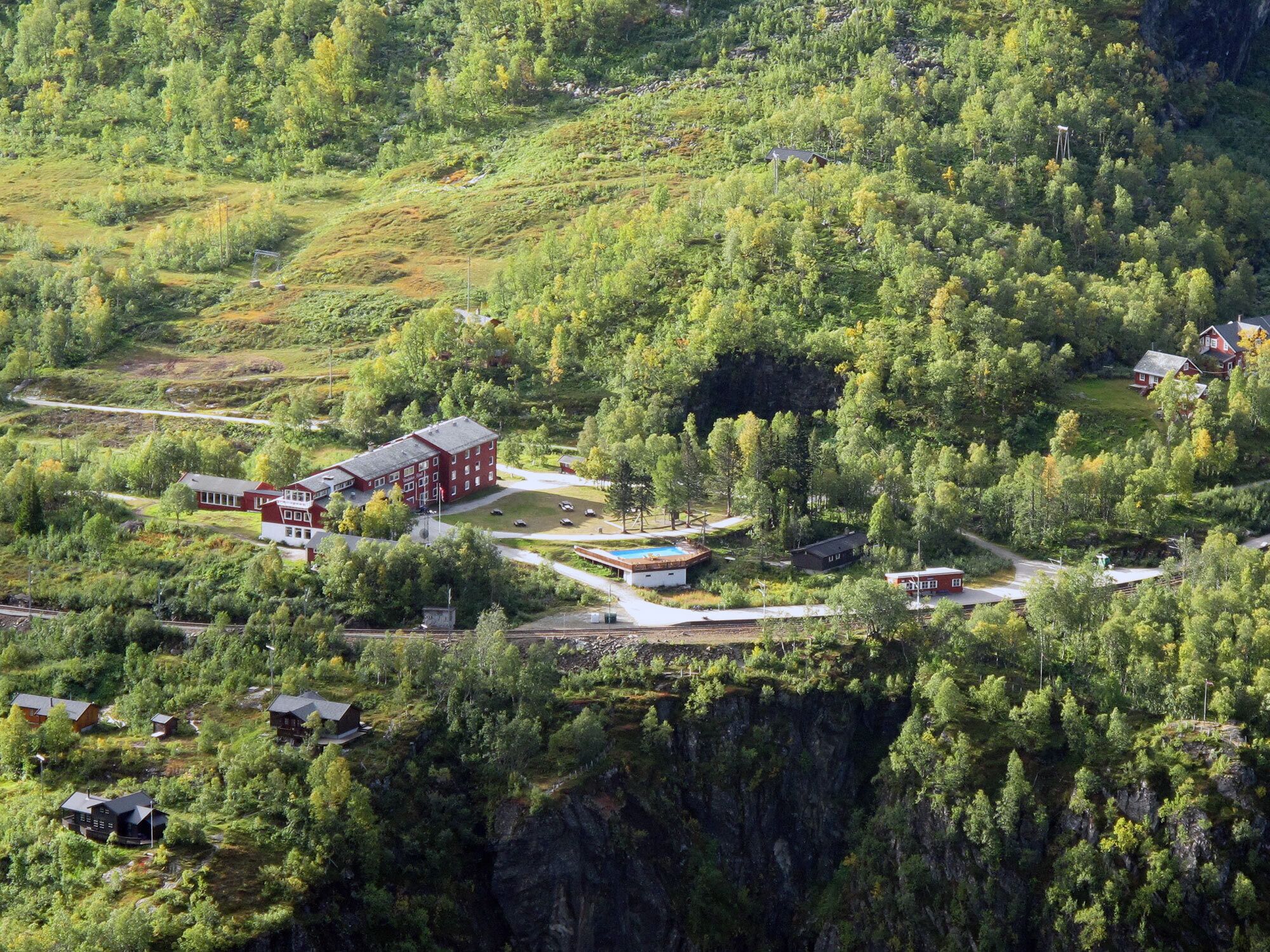 A remote, forested area with a cluster of buildings, including a large red building and several smaller structures, surrounded by dense trees and a steep cliff.