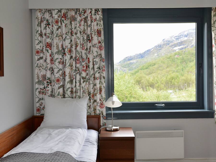 Una camera da letto con un letto, un comodino, una lampada, una finestra vista montagne e tende a fiori.