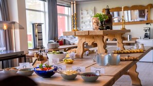 A buffet setup with various food items and labels, a wooden table, and a window with curtains in the background. - Vatnahalsen Høyfjellshotell (Aurland)
